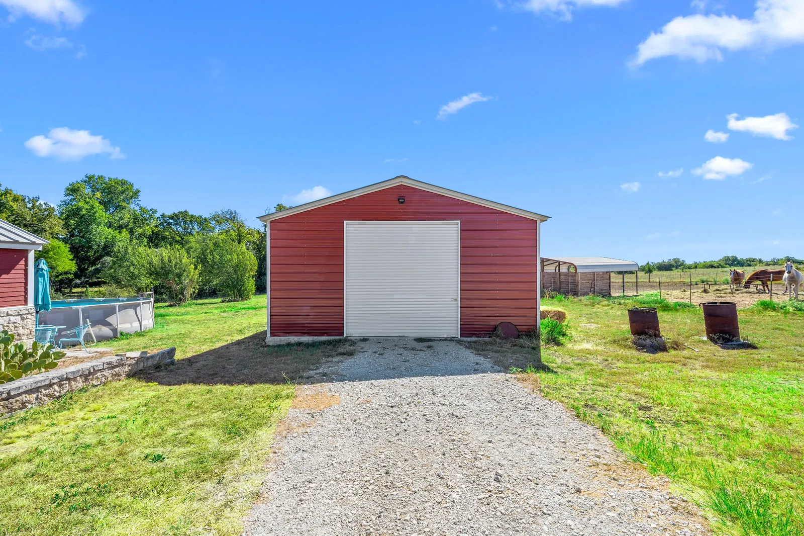 Wide-angle view of a large red metal shop with roll-up doors beside a rural farmhouse.