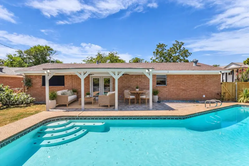 Expansive backyard featuring a swimming pool with integrated steps, a covered patio with a paved surface, and a brick exterior, virtually staged