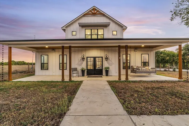 Modern farmhouse exterior featuring vertical siding, a deep wrap-around covered porch with wood support columns, dual-entry glass panel doors, and architectural gable detailing